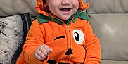 George is registered to the contest to win money with this photo: baby, child, costume, couch, cozy, cute, cute_outfit, fall, festive, happy, holiday, hood, indoors, orange_hoodie, person, portrait, pumpkin_costume, seated, smiling, striped_socks