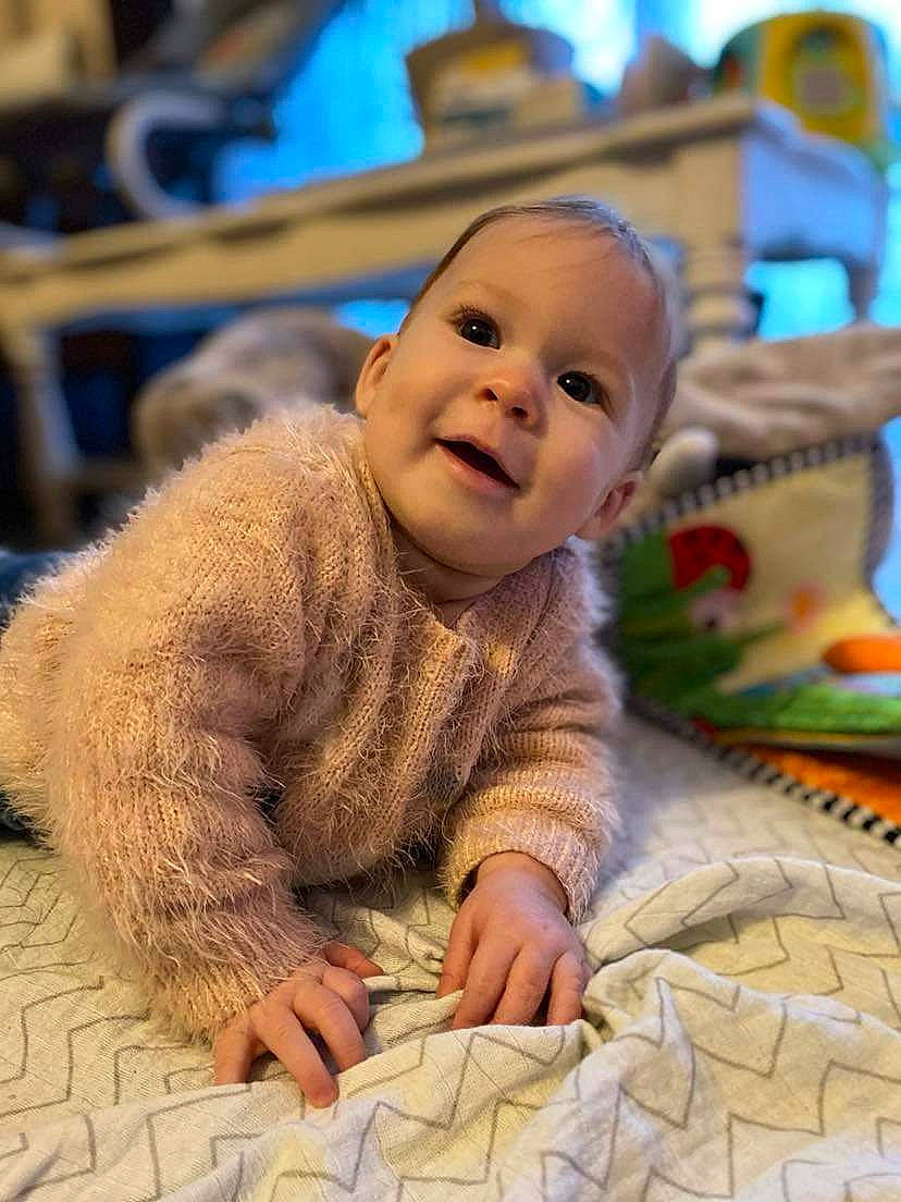 Lola participe au concours pour gagner de l'argent avec cette photo : baby, baby_toddler_clothing, carpet, cheek, child, comfort, facial_expression, floor, flooring, fun, fur, happy, headwear, leisure, linens, person, room, sitting, skin, smile