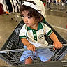 blue_shorts, casual_clothing, child, clothing_store, curious, curly_hair, footwearless, green_trim, holding, indoor, person, polo_shirt, portrait, retail, shopping, shopping_cart, store_aisle, toddler, white_cap, young_child