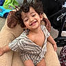 baby_toys, casual, child, comfortable, curly_hair, cute, expression, happy, indoor, person, playful, plush_chair, portrait, seated, smiling, striped_clothing, stroller, toddler, toy_car, young_child