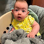 Addison is registered to the contest to win money with this photo: baby, child, laundry_basket, towel, yellow_clothing, daisy_pattern, infant, surprised_expression, indoor, soft_texture, cute, small_hand, seated, plush, comfort, home, portrait, person, face, curious