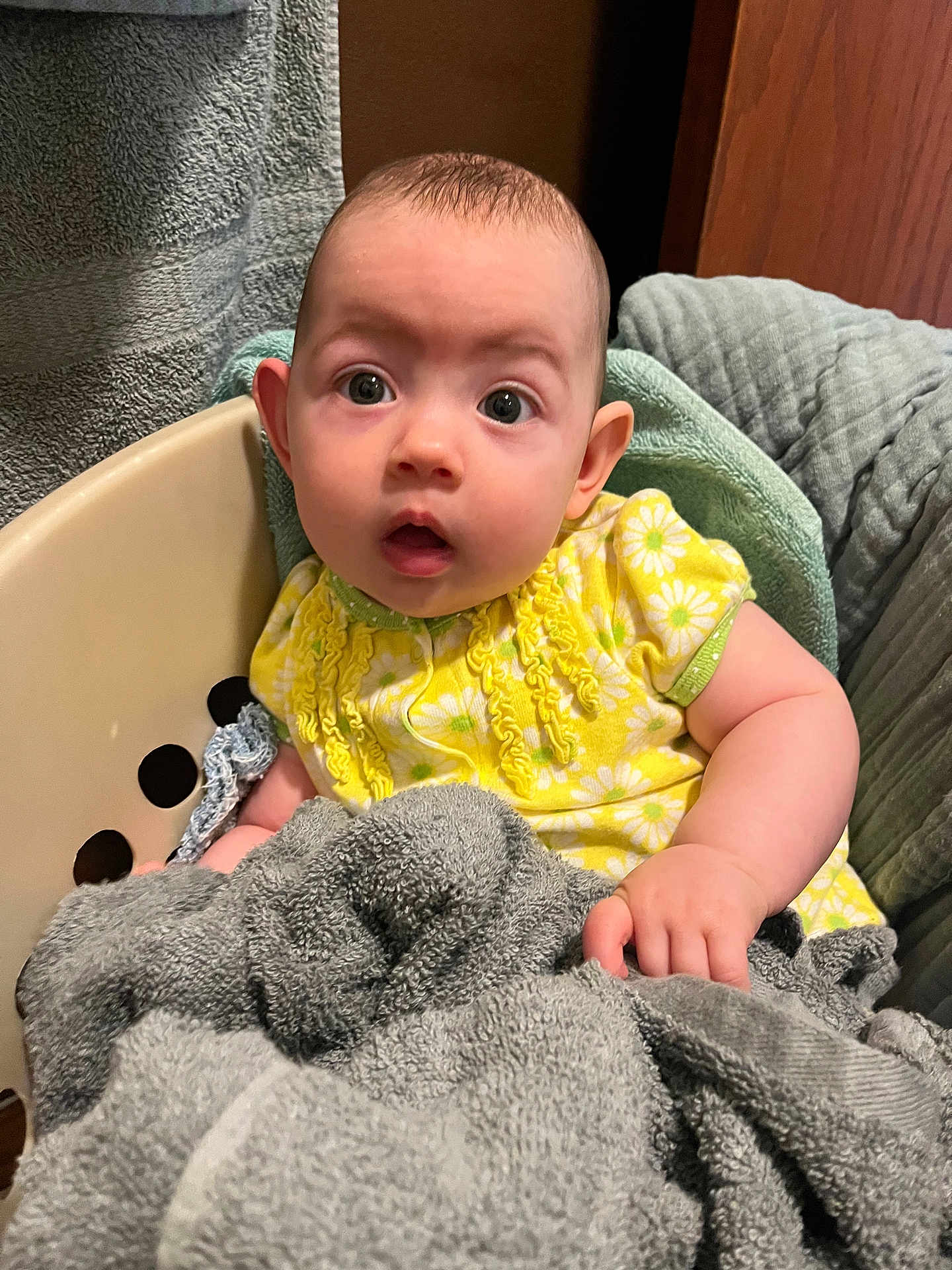Addison is registered to the contest to win money with this photo: baby, child, laundry_basket, towel, yellow_clothing, daisy_pattern, infant, surprised_expression, indoor, soft_texture, cute, small_hand, seated, plush, comfort, home, portrait, person, face, curious