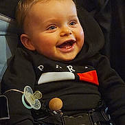 Julianno participe au concours pour gagner de l'argent avec cette photo : accessory, baby, belt, black_clothing, blond_hair, blue_eyes, candid, cheeks, child, hand, happy, harness, hoodie, indoors, infant, pacifier, portrait, seat, smiling, teeth