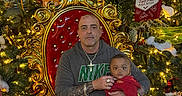 Victor is registered to the contest to win money with this photo: man, baby, chair, christmas_tree, holiday_decorations, lights, ornate_chair, red, gold, festive, indoor, person, child, family, seated, celebration, cross_necklace, sweatshirt, decor, smiling