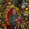 man, baby, chair, christmas_tree, holiday_decorations, lights, ornate_chair, red, gold, festive, indoor, person, child, family, seated, celebration, cross_necklace, sweatshirt, decor, smiling