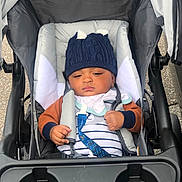 Victor is registered to the contest to win money with this photo: baby, stroller, hat, knit_hat, child, infant, seat_belt, outdoor, clothing, striped_shirt, jacket, face, person, hands, headwear, safety, baby_bib, wheel, transport, cute
