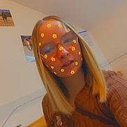 Lana participe au concours pour gagner de l'argent avec cette photo : person, indoor, glasses, blonde_hair, flower_filter, bed_frame, brown_sweater, seated, portrait, casual, wall_photos, cozy, young_person, smile, head_and_shoulders, light, home, decor, face_filter, glasses_reflection