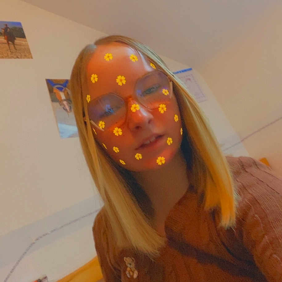 Lana participe au concours pour gagner de l'argent avec cette photo : person, indoor, glasses, blonde_hair, flower_filter, bed_frame, brown_sweater, seated, portrait, casual, wall_photos, cozy, young_person, smile, head_and_shoulders, light, home, decor, face_filter, glasses_reflection