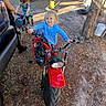 child, children, dirt_bike, bicycle, outdoor, backyard, tree, dirt, sunlight, play, toy, person, grass, smile, blue_shirt, red_bike, casual, daytime, happy, nature