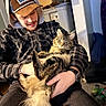 man, cat, indoor, plaid_jacket, hat, cap, tabby_cat, fluffy_cat, holding, sitting, casual, facial_hair, watch, cardboard_box, clutter, container, carpet, relaxed, pet, cozy