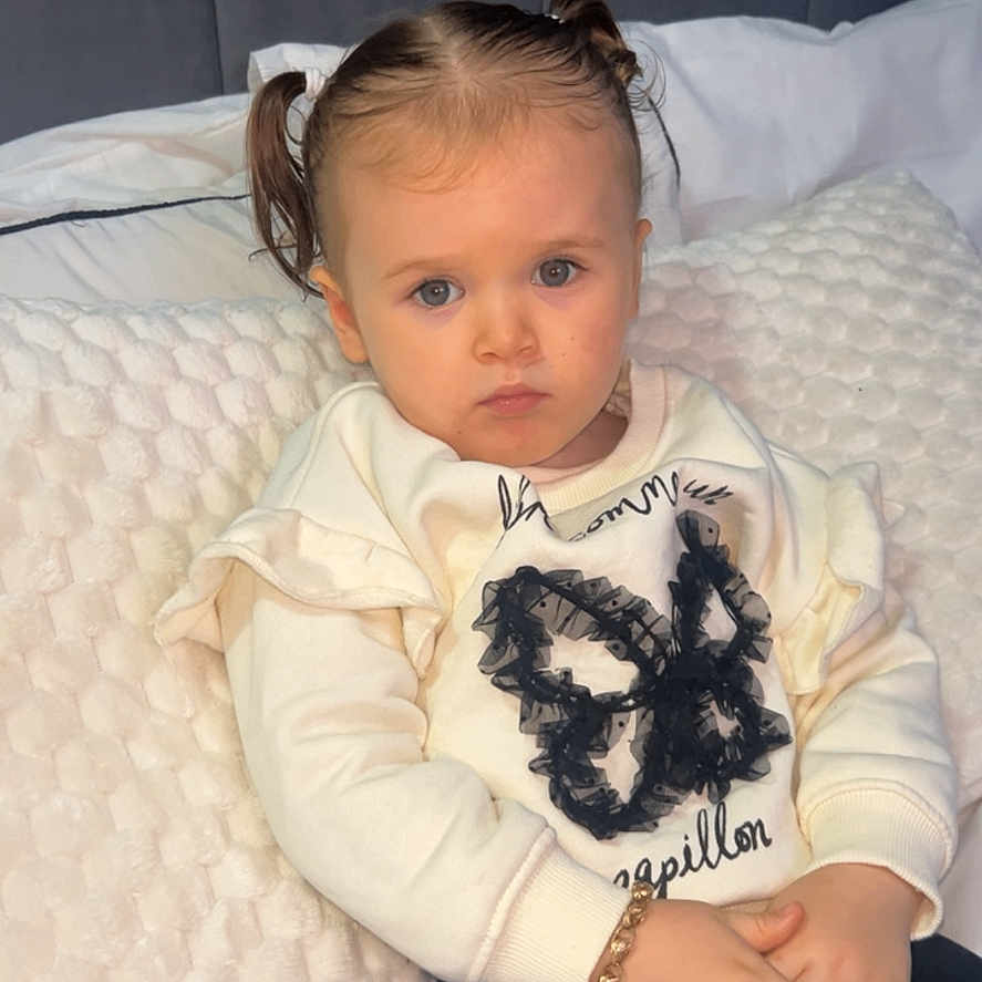 Zylah is registered to the contest to win money with this photo: bed, bracelet, butterfly, casual, child, cozy, cute, fashion, hands, indoors, person, pigtails, pillow, portrait, serious, sitting, sweater, toddler, white, young