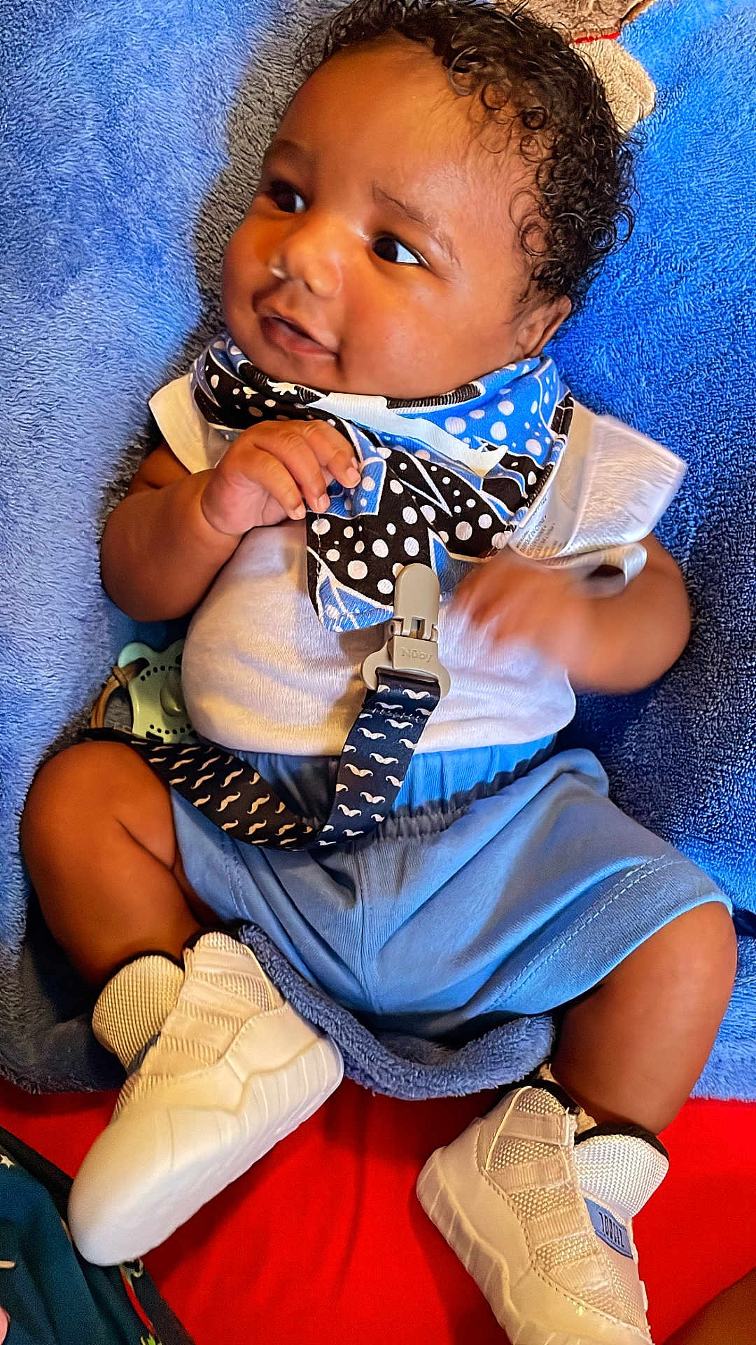 Hayden joined the competition — help win amazing prizes! baby, infant, child, blue_shorts, white_shoes, bib, blanket, pacifier_clip, curly_hair, cute, soft_texture, lying_down, casual_clothing, indoor, relaxed, close_up, person, smiling, skin, footwear