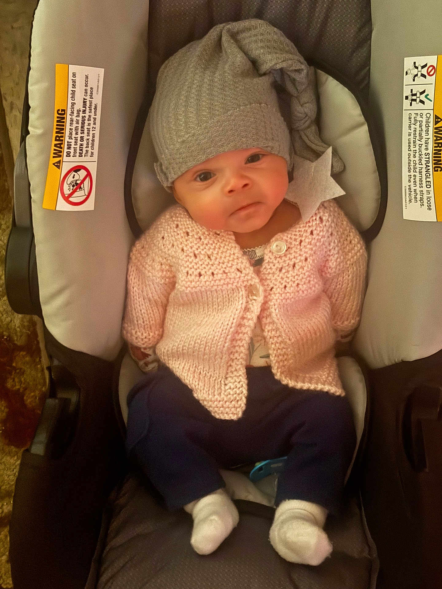 Chloee is registered to the contest to win money with this photo: baby, infant, child, car_seat, hat, knitted_sweater, pink_clothing, gray_hat, star, white_socks, blue_pants, indoors, cute, young_child, seated, car_seat_warning_label, portrait, cozy, warm_clothing, looking_at_camera