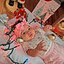 baby, infant, pink_bow, headband, christmas_lights, ornaments, holiday_decor, santa_figurine, wooden_floor, blanket, festive, cute, toddler, toy, red_pants, vest, smiling, celebration, indoors, holiday