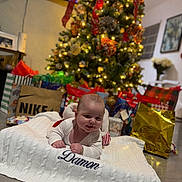 Damon Jiménez Romeu is registered to the contest to win money with this photo: baby, blanket, christmas_tree, gifts, holiday, festive, smiling, indoors, floor, decorations, lights, red_ribbon, cozy, happy, child, person, home, celebration, wrapped_presents, knitted