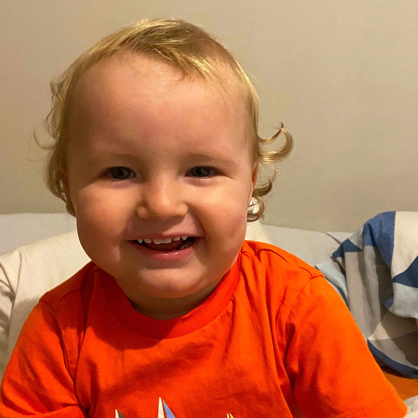 Baptiste a rejoint le concours — aidez-le/la à gagner de superbes lots ! bed, blanket, blond_hair, bright_color, casual, child, clothing, cozy, curly_hair, cute, face, happy, indoors, orange_tshirt, person, portrait, sitting, smile, toddler, young_child