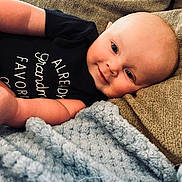 Oden joined the competition — help win amazing prizes! baby, smiling, blanket, black_shirt, text_on_clothing, blue_blanket, lying_down, face, infant, cute, happy, skin, eyes, head, soft_texture, comfort, cozy, closeup, person, indoors