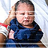 newborn, baby, infant, child, portrait, face, eyes_open, looking_at_camera, blue_clothing, swaddled, blanket, adult_hand, hospital_bracelet, indoor, photograph, smartphone_screen, website, watermark, close_up, sitting
