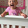 baby, crib, pink_onesie, smiling, laughing, child, indoor, furniture, baby_clothing, hands, face, head, person, bedroom, quilt, blanket, wood, infant, happy, standing