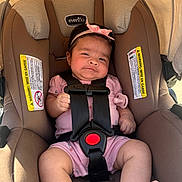 Valentina is registered to the contest to win money with this photo: baby, brown_car_seat, canopy, car_seat, child, comfortable, cute, expression, headband, indoors, infant, pacifier, person, pink_bow, pink_outfit, seatbelt, sitting, straps, sunlight, warning_labels