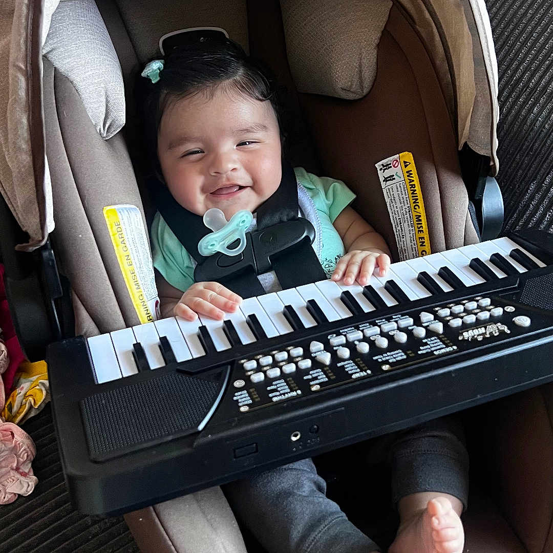 Valentina is registered to the contest to win money with this photo: baby, car_seat, child, clothing, cushion, feet, footwear, hands, happy, indoor, infant, keyboard, music, pacifier, plush_toy, portrait, seat_belt, small_keyboard, smiling, toy