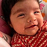 baby, smiling, face, child, red_clothing, heart_pattern, blanket, soft_toys, indoors, cute, portrait, happy, young_child, cozy, infant, head, expression, warm_colors, resting, closeup