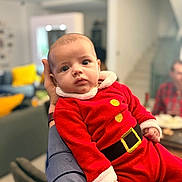 Lorenzo participe au concours pour gagner de l'argent avec cette photo : baby, child, person, red_clothing, santa_outfit, indoor, adult_arm, face, wide_eyes, plush_fabric, hand, furniture, living_room, blurred_background, couch, yellow_pillow, portrait, cute, holiday, cozy