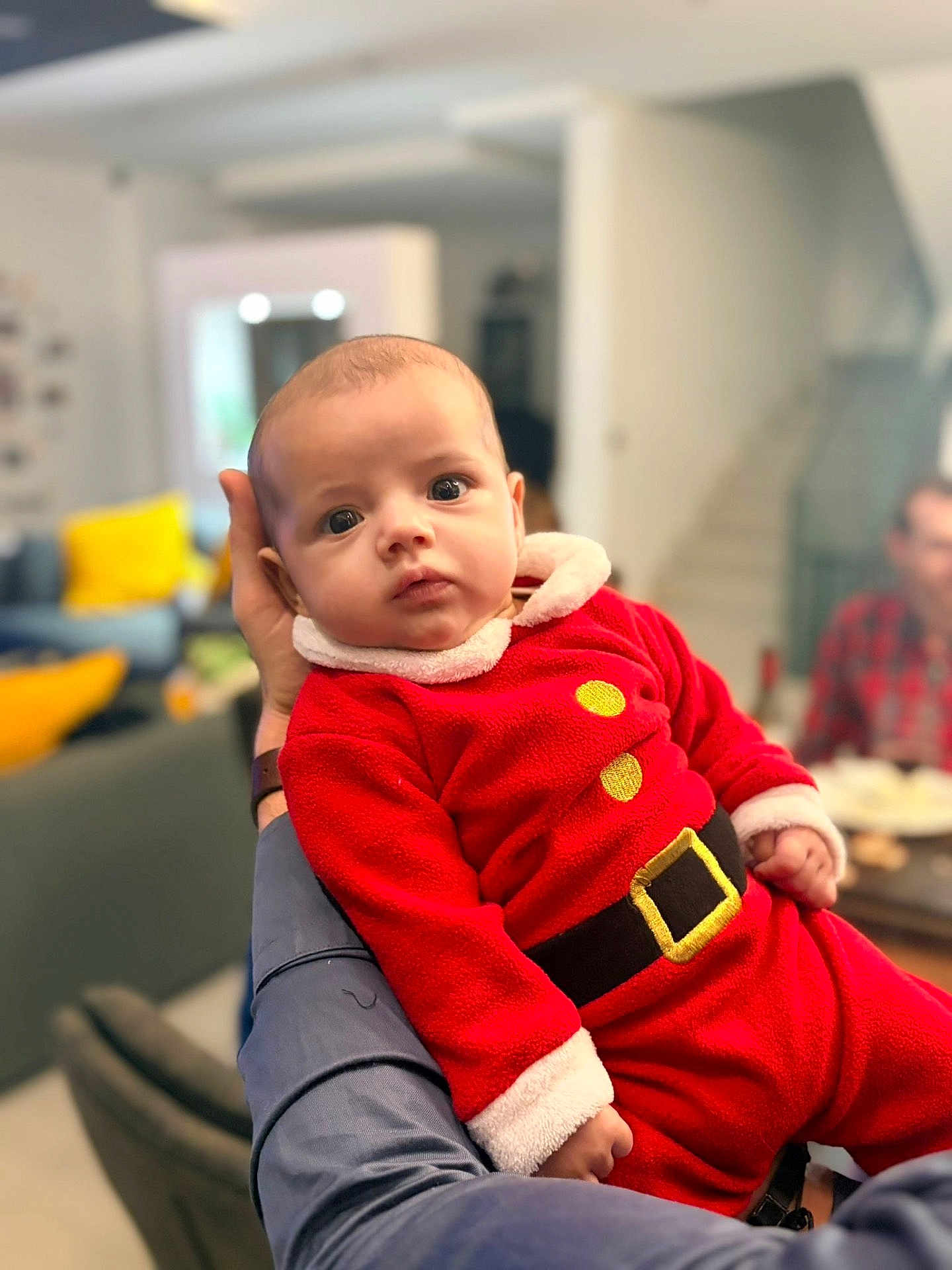 Lorenzo participe au concours pour gagner de l'argent avec cette photo : baby, child, person, red_clothing, santa_outfit, indoor, adult_arm, face, wide_eyes, plush_fabric, hand, furniture, living_room, blurred_background, couch, yellow_pillow, portrait, cute, holiday, cozy