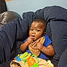 baby, infant, child, portrait, sitting, couch, blanket, blue_cushions, toy, rattle, teether, bib, blue_shirt, brown_skin, curious, hand_in_mouth, indoors, home, furniture, smiling