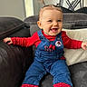 toddler, child, smiling, sitting, couch, overalls, red_clothing, blue_clothing, pigtails, baby, indoor, happy, face, person, sock, furniture, dartboard, home, cute, portrait