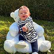 Maëlle a rejoint le concours — aidez-le/la à gagner de superbes lots ! baby, child, teddy_bear, plush, outdoor, grass, hedge, sunlight, striped_shirt, footwear, happy, smiling, sitting, nature, greenery, cute, young_child, playful, portrait, daylight