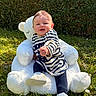 baby, child, teddy_bear, plush, outdoor, grass, hedge, sunlight, striped_shirt, footwear, happy, smiling, sitting, nature, greenery, cute, young_child, playful, portrait, daylight