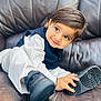 toddler, child, smile, shoe, black_shoe, white_clothing, navy_vest, leather_couch, indoor, cute, portrait, happy, young_child, hand, footwear, sitting, face, big_eyes, curly_hair, playful