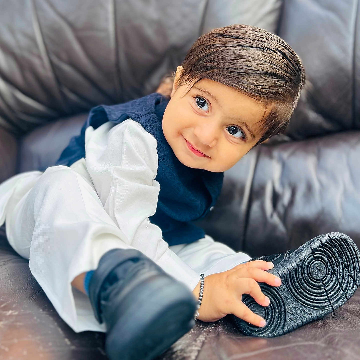 Gurupdesh joined the competition — help win amazing prizes! big_eyes, black_shoe, child, curly_hair, cute, face, footwear, hand, happy, indoor, leather_couch, navy_vest, playful, portrait, shoe, sitting, smile, toddler, white_clothing, young_child
