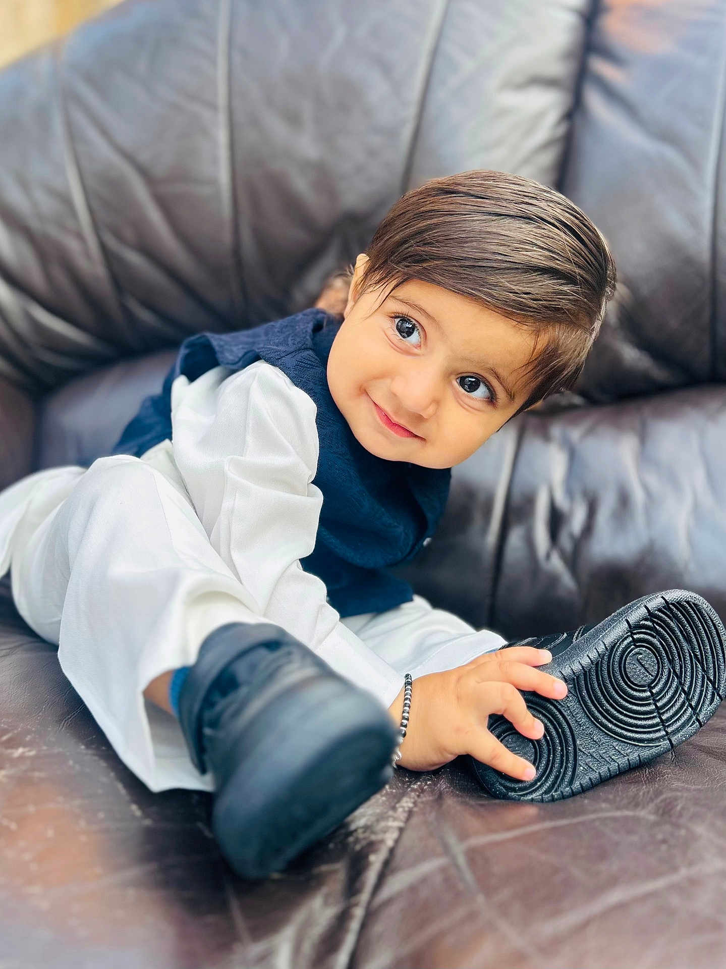 Gurupdesh joined the competition — help win amazing prizes! toddler, child, smile, shoe, black_shoe, white_clothing, navy_vest, leather_couch, indoor, cute, portrait, happy, young_child, hand, footwear, sitting, face, big_eyes, curly_hair, playful