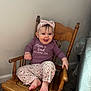 baby, child, smiling, rocking_chair, wooden_chair, headband, floral_pattern, purple_clothing, long_sleeve, pants, barefoot, indoor, carpet, happy, cute, person, sitting, infant, portrait, home
