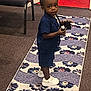 toddler, child, indoor, carpet, floral_pattern, navy_blue_clothing, shorts, shirt, white_sneakers, curious_expression, holding_object, runner, chair, carpeted_floor, earring, person, standing, looking_back, black_hair, small_object