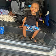 Omarius is registered to the contest to win money with this photo: child, toddler, smiling, sitting, car, car_seat, shorts, black_shirt, footwear, red_bull_can, blanket, cooler, vehicle_interior, person, happy, casual, curious, candid, indoor, relaxed