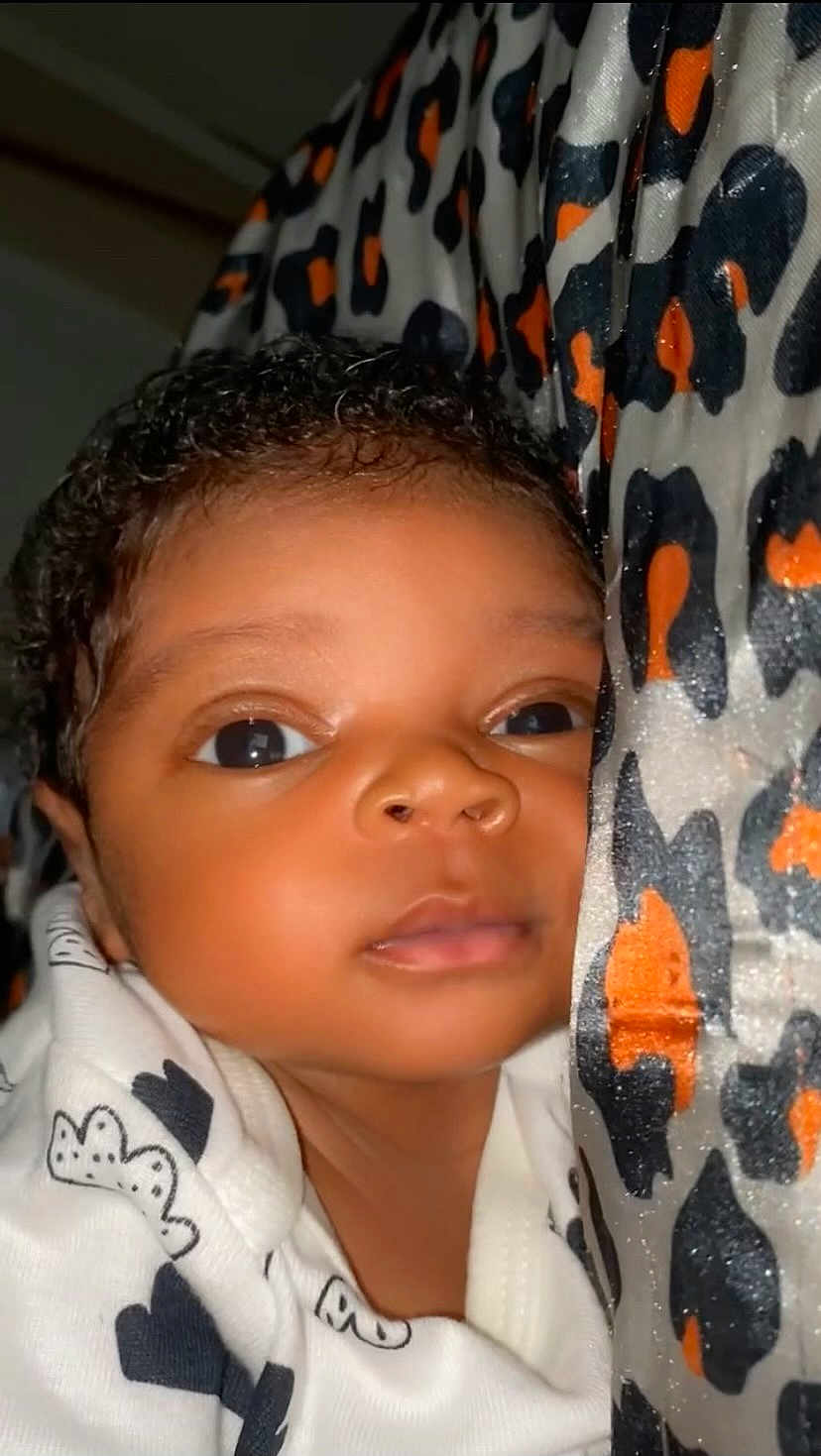 Omarius is registered to the contest to win money with this photo: baby, infant, face, close_up, curly_hair, blanket, leopard_print, pattern, white_clothing, black_pattern, eyes_open, skin, portrait, young_child, cute, resting, indoors, soft_light, cozy, newborn