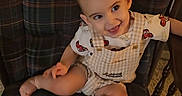 Raylan is registered to the contest to win money with this photo: baby, child, plaid_chair, armchair, smiling, happy, indoor, wooden_marker, milestone, cute_outfit, shorts, shirt, sitting, infant, person, furniture, plaid_pattern, young_child, portrait, cozy