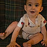 baby, child, plaid_couch, overalls, white_shirt, red_cars, smiling, curious, sitting, indoor, hand, foot, round_object, young_child, portrait, cute, face, person, furniture, casual_clothing