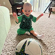 Raylan joined the competition — help win amazing prizes! toddler, child, soccer_ball, carpet, indoor, green_clothing, smiling, sitting, furniture, rocking_chair, window, natural_light, playful, home, sock, table, curtains, floor, person, toy