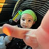 baby, child, car_seat, green_hat, reaching_hand, tongue_out, striped_canopy, car_interior, sunlight, curious_expression, infant, seatbelt, clothing, person, portrait, cute, indoors, seat, safety, closeup