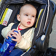 Raylan is registered to the contest to win money with this photo: toddler, child, stroller, water_bottle, outdoor, seatbelt, clothing, baby, person, sunlight, sidewalk, nature, trees, curious, expression, safety, infant, head, face, hands