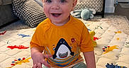 Liam is registered to the contest to win money with this photo: baby, child, smiling, sitting, orange_shirt, shark_graphic, striped_shorts, playmat, animal_print, living_room, couch, pillow, shelf, toys, carpet, happy, indoor, person, hair_style, cute