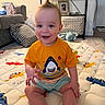 baby, child, smiling, sitting, orange_shirt, shark_graphic, striped_shorts, playmat, animal_print, living_room, couch, pillow, shelf, toys, carpet, happy, indoor, person, hair_style, cute