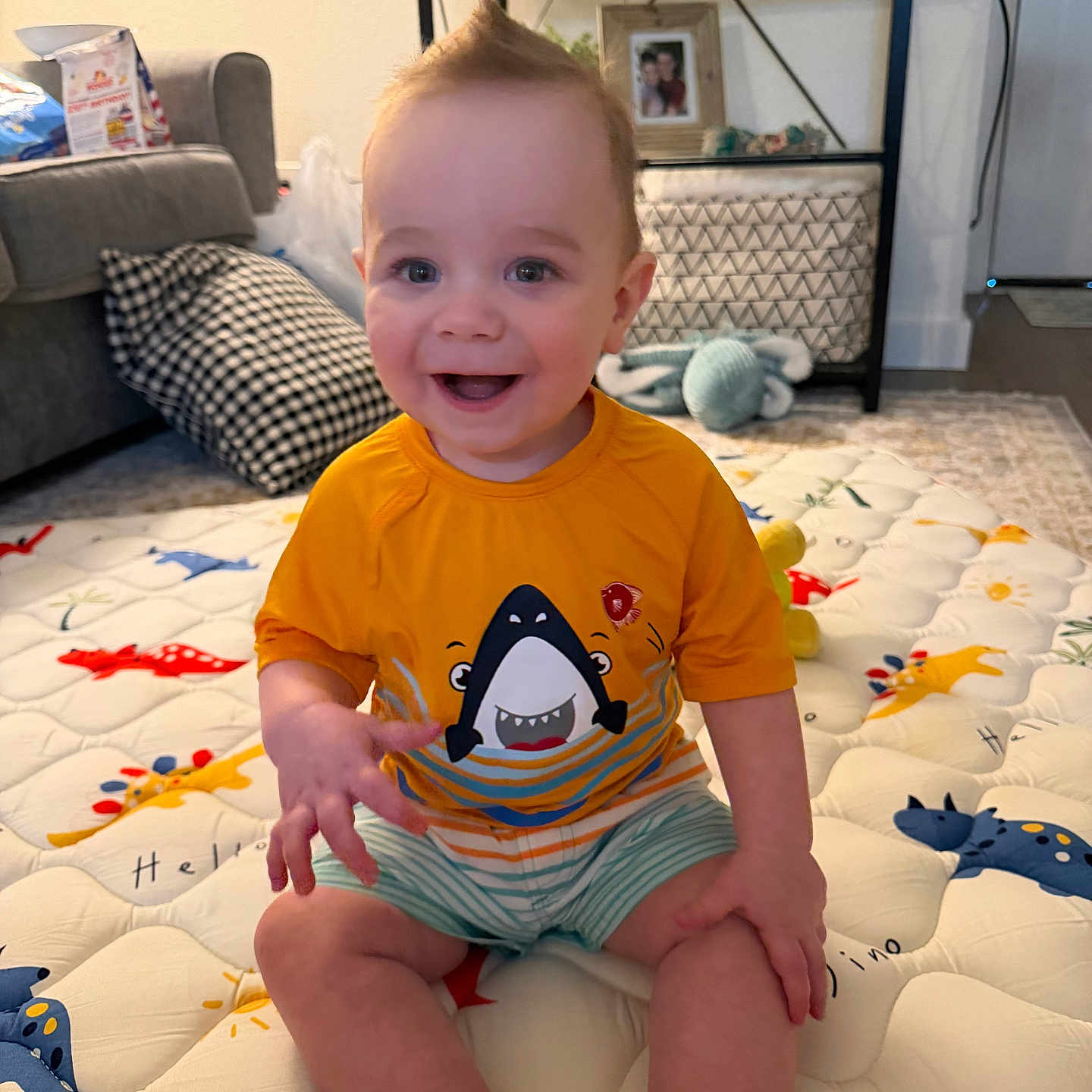 Liam is registered to the contest to win money with this photo: baby, child, smiling, sitting, orange_shirt, shark_graphic, striped_shorts, playmat, animal_print, living_room, couch, pillow, shelf, toys, carpet, happy, indoor, person, hair_style, cute
