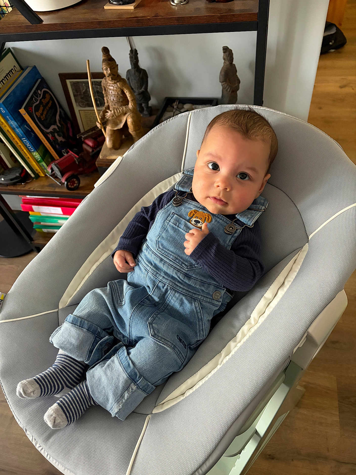 Theo a rejoint le concours — aidez-le/la à gagner de superbes lots ! baby, infant, denim_overalls, striped_socks, baby_bouncer, baby_seat, bookshelf, books, wooden_statue, toy_car, indoor, hardwood_floor, face, big_eyes, curious_expression, hand, cozy, portrait, clothing, cute