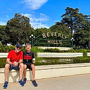 James is registered to the contest to win money with this photo: adult, boy, smiling, sitting, fountain, beverly_hills_sign, outdoor, daytime, trees, greenery, sky, casual_clothing, shorts, sneakers, glasses, cap, coffee_cup, tourist, landmark, urban