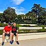 adult, boy, smiling, sitting, fountain, beverly_hills_sign, outdoor, daytime, trees, greenery, sky, casual_clothing, shorts, sneakers, glasses, cap, coffee_cup, tourist, landmark, urban